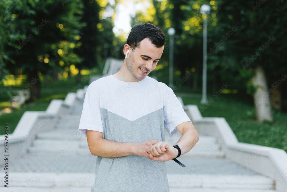Young smiling man setting up his smart watch of fitness band before work out or checking it out after that in park or outdoors.