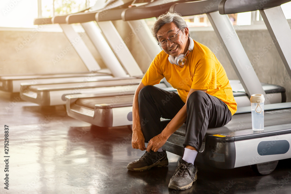 Senior asian sport man tying shoelaces on treadmill in fitness gym ready walking with headphone and water bottle . elder male exercising , workout, training ,healthy ,Retirement ,older, looking camera Senior asian sport man tying shoelaces on treadmill in fitness gym ready walking with headphone and water bottle . elder male exercising , workout, training ,healthy ,Retirement ,older, looking camera