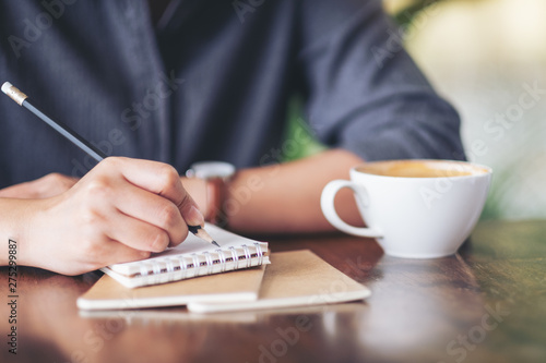 Closeup image of a woman writing on blank notebook with coffee cup on table in cafe