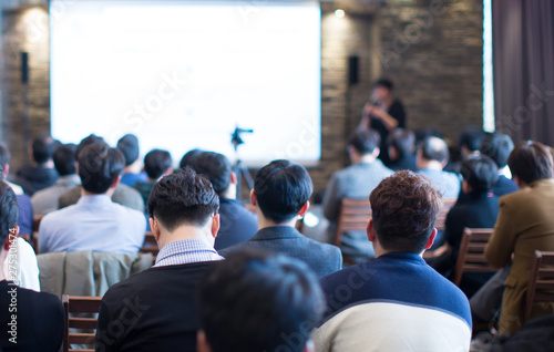 "Speaker giving a talk at a business conference. Audience in hall with presenter next to presentation screen. Executive giving speech during business and entrepreneur seminar. "
