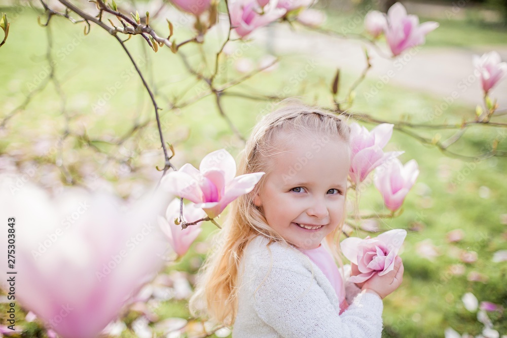 Fototapeta premium Little cute blonde girl 3 years old plays in a park near a blossoming magnolia.