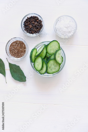 Fototapeta Naklejka Na Ścianę i Meble -  Flat lay composition of fresh cucumber sliced in a glass bowl and the necessary spices for pickling(fermenting)- dill seeds, pepper, salt on a white wooden table.Top view, copy space for text.