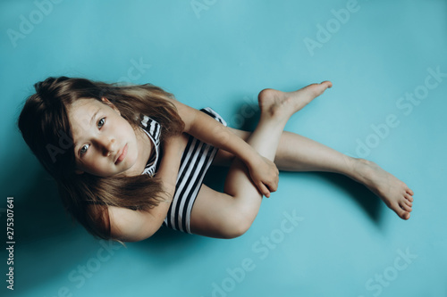 Girl sitting on plain blue background looking at camera. Child glancing up top view. Simple wallpaper  background. Serious and sad facial expression. Kid in striped dress with legs crossed