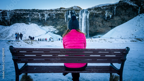 A girl in pink jacket sits on the bench and looking tourists in front of the frozen Seljalandsfoss waterfall in wintertime. It is one of most popular place in South Coast Iceland.