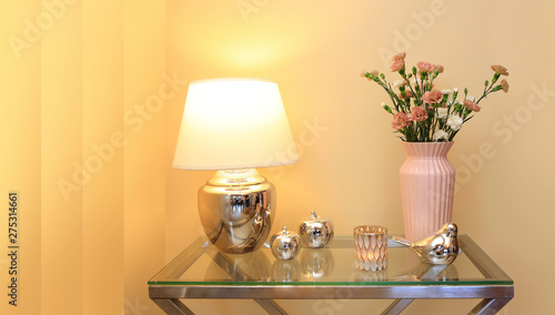 Lampka nocna i kwiaty w wazonie na szklanym stoliku i złotych barwach. 
