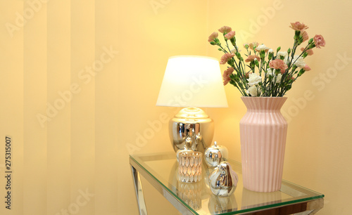 Lampka nocna i kwiaty w wazonie na szklanym stoliku i złotych barwach.