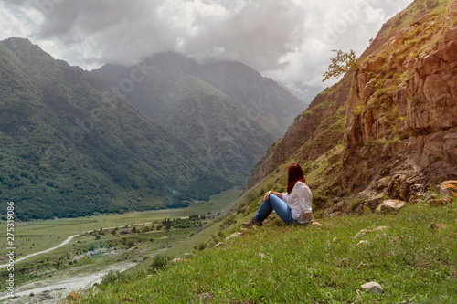 a woman with red hair in a white blouse and jeans sits against a mountain landscape, toned