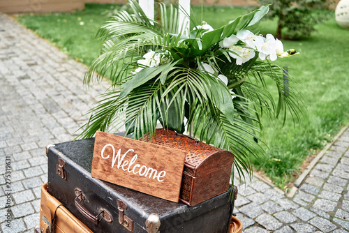 Wedding decorations with welcome sign board, flowers, pulm leaves and vintage suitcases in garden, copy space photo