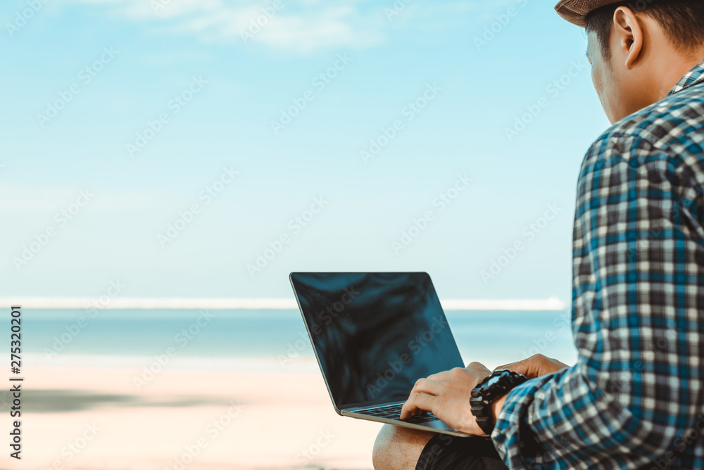 freelancer young man in a shirt and shorts and sneakers working on laptop while sitting on the beach, sending mail,