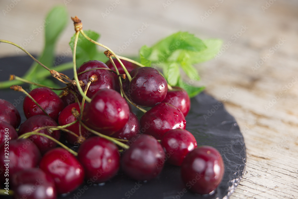 Fresh cherries on a black plate of wet slate with sprig of fresh green mint on light wooden table in selective focus for copy space Fresh cherries on a black plate of wet slate with sprig of fresh green mint on light wooden table in selective focus for copy space
