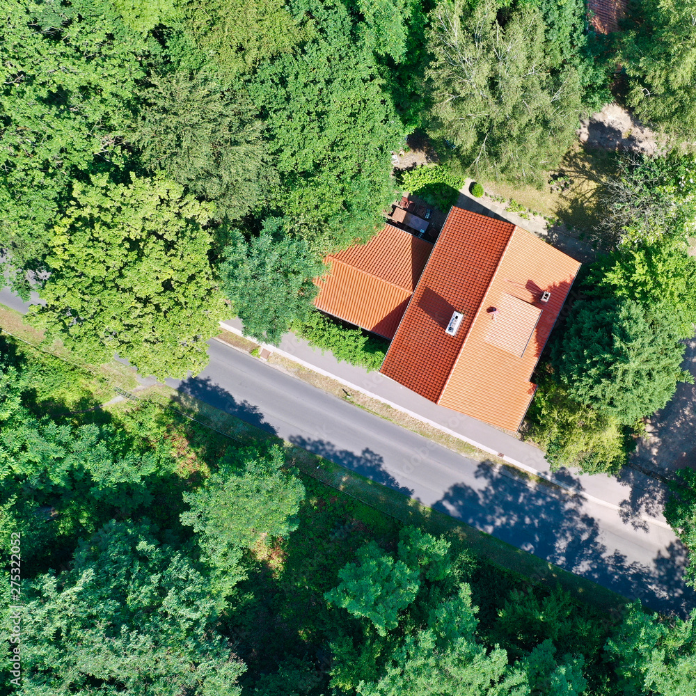 Vertical aerial view of a house with a saddle roof and red roof tiles in a wooded area next to an asphalted grey country road. Vertical aerial view of a house with a saddle roof and red roof tiles in a wooded area next to an asphalted grey country road.