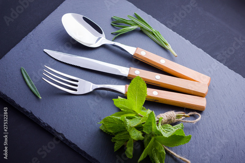 Cutlery set. Knife, spoon, fork on slate stone. Сutlery with rosemary and mint. Black background. Copy space. Top view
