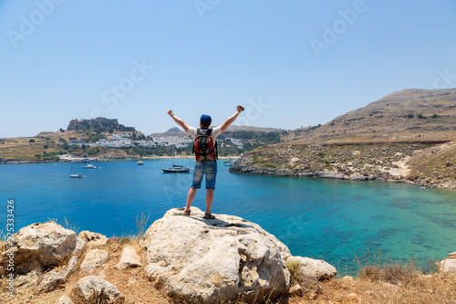 Young man with backpack standing with raised hands on top of a mountain, castle, sea and enjoying view. Greece, Lindos.