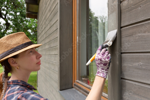 Woman worker painting wooden house exterior wall with paintbrush and wood protective color