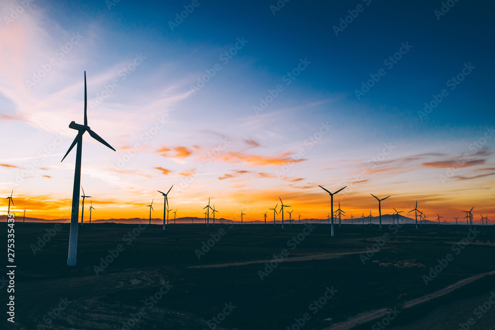 Obraz premium Silhouette of windmills station with propellers generating alternative clean green power from eco resources in rural agri environment against blue sunset sky. Farm of turbines producing electricity