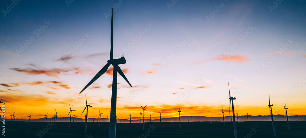 Obraz premium Silhouette of windmills station with propellers generating alternative clean green power from eco resources in rural agri environment against blue sunset sky. Farm of turbines producing electricity