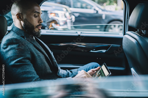 Side view of African American male broker checking information on trade website connected to 4g wireless during business trip in car,dark skinned director holding digital tablet for communication