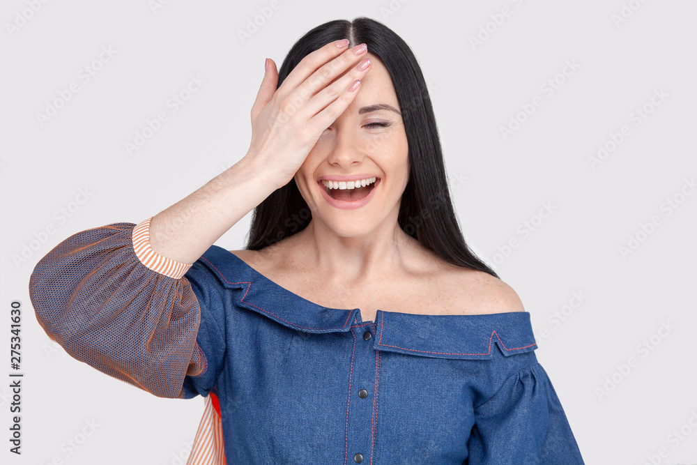 Cheerful pretty charming woman in jeans dress with dark hair smiling happily, having fun indoors, closing one eye with hand. Pretty girl looking at camera with joyful smile Cheerful pretty charming woman in jeans dress with dark hair smiling happily, having fun indoors, closing one eye with hand. Pretty girl looking at camera with joyful smile