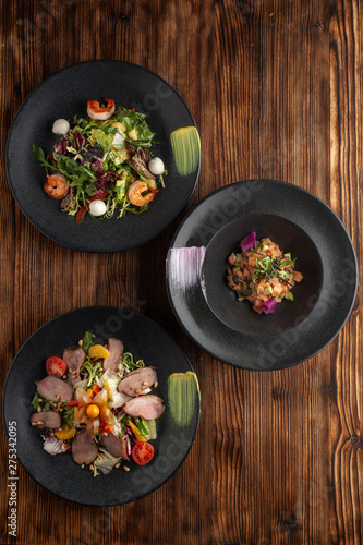 Variety of fancy appetizers on a minimalist wooden background: raw salmon tartare with avocado, green salad with prawns, duck breast salad with orange dressing