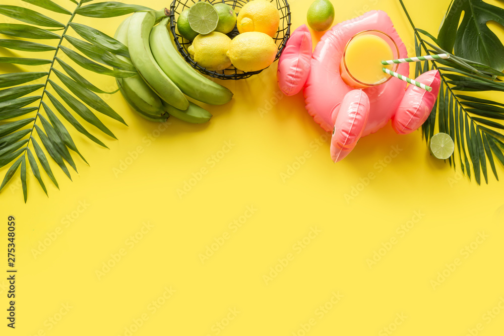 Tropical frame of fruits, banana, lime, leaves palms, orange juice in inflatable pink flamingo on punchy pastel yellow background.