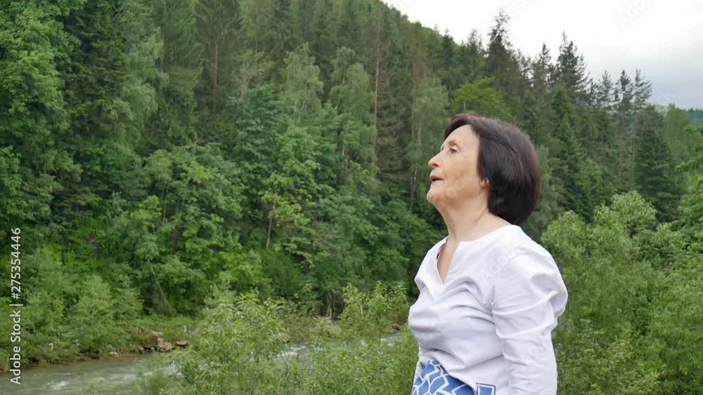 Senior woman with short dark hair doing a stretching exercise for relaxation in the morning outside over landscape of forest and mountains Senior woman with short dark hair doing a stretching exercise for relaxation in the morning outside over landscape of forest and mountains