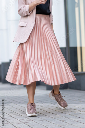 Model posing in a peach skirt, pleated, sneakers and jacket. Coral or pink color. On the street in the afternoon.