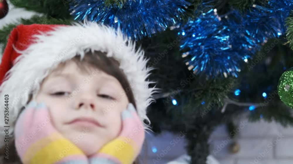 Kid with Santa Claus hat near the Christmas tree. Portrait of a cute little girl waiting for christmas. Christmas baby. Child waiting for the Christmas holiday. Kid with Santa Claus hat near the Christmas tree. Portrait of a cute little girl waiting for christmas. Christmas baby. Child waiting for the Christmas holiday.