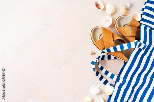 Striped beach bag with casual trendy leather sandals with crisscross details, shells on pink pastel background. Vacation holidays layout.