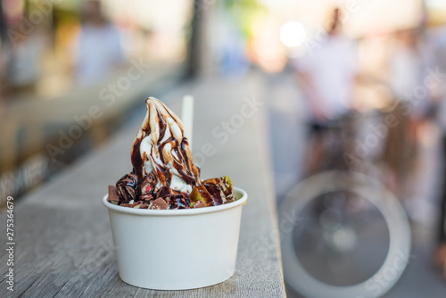 Closeup outdoor view at White Frozen Yogurt serve with topping with fruit and chocolate in paper white cup, on wooden counter table on street, and blur background people and bicycle in summer season.  photo