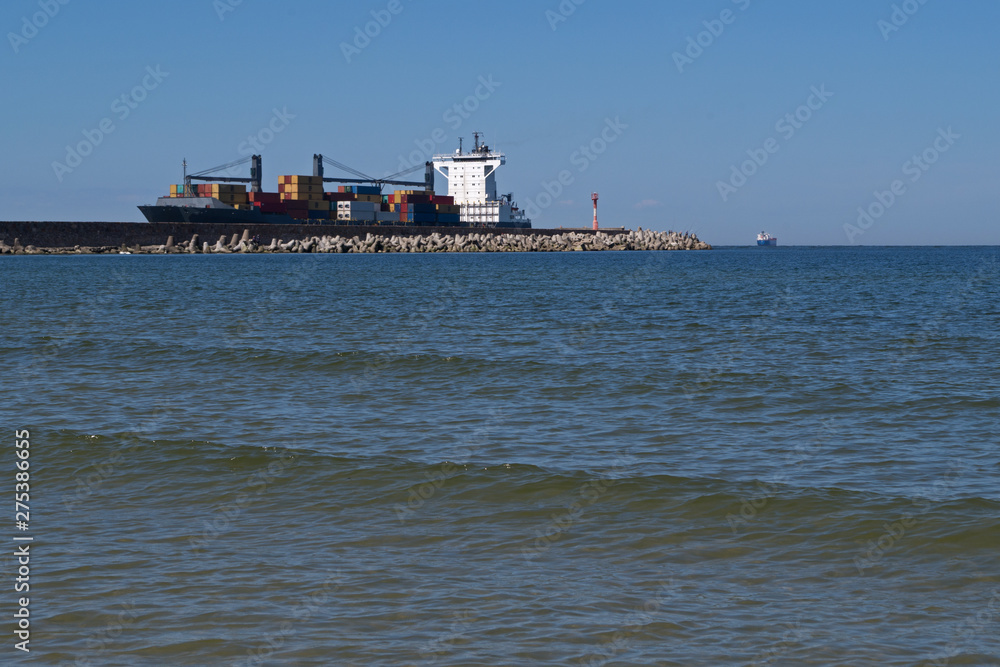 Blue hulled cargo ship loaded with shipping containers of various colors enters the coastal Bay past a pier with a lighthouse on the edge and concrete breakwaters on the side against a clear blue sky