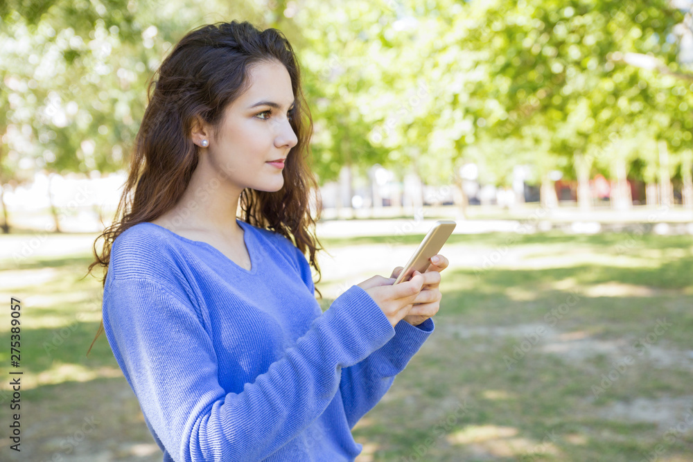 Pensive young woman using modern gadget while strolling in city park. Content girl with wavy hair thinking about text of message. Online communication concept Pensive young woman using modern gadget while strolling in city park. Content girl with wavy hair thinking about text of message. Online communication concept