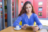Young woman using tablet in sidewalk cafe. Thoughtful attractive girl with wavy hair using mobile internet while drinking coffee outdoors. City break concept