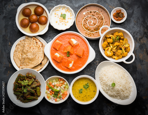 Assorted Indian food like paneer butter masala  dal  roti  rice  sabji  gulab jamun and bound raita served in bowls over moody background  selective focus