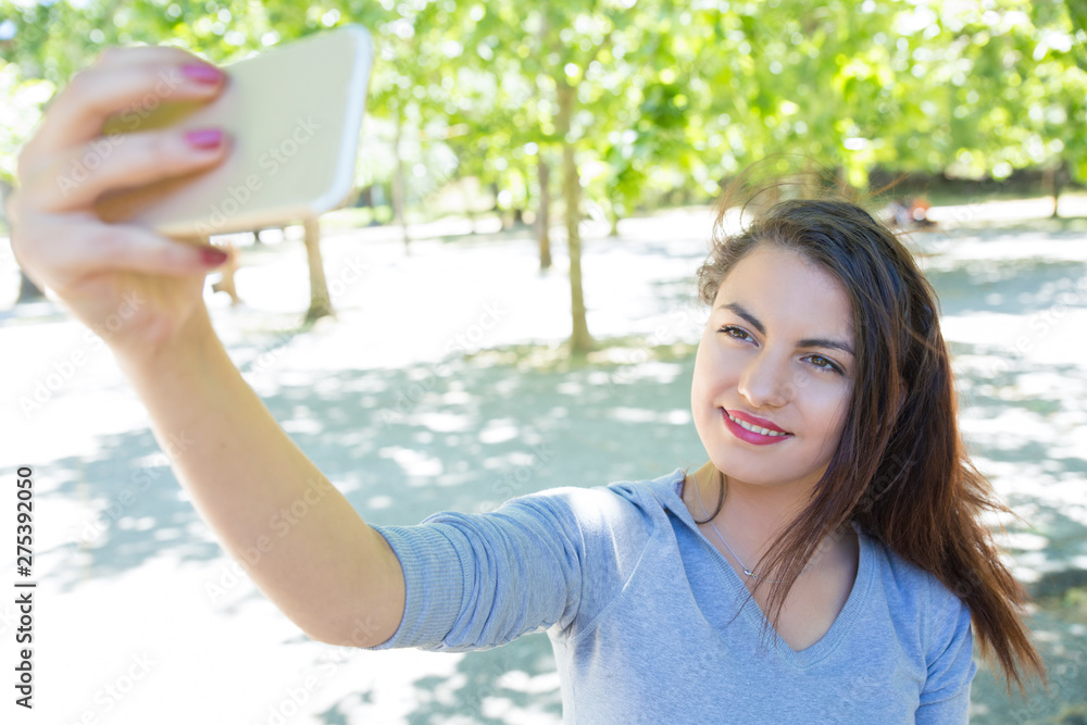 Happy pretty young lady taking selfie photo in park. Beautiful lady wearing hoodie and posing with green trees in background. Selfie and nature concept. Front view. Happy pretty young lady taking selfie photo in park. Beautiful lady wearing hoodie and posing with green trees in background. Selfie and nature concept. Front view.