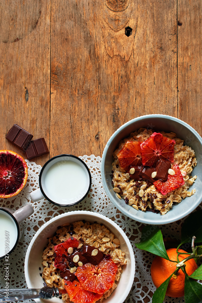 Two bowls of porridge with red oranges and chocolate and fresh milk over wooden table. Flat lay with space for text.