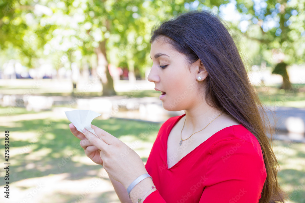 Pretty young woman holding tissue and sneezing in park. Beautiful lady wearing red blouse and standing with green lawn and trees in background. Cold and nature concept. Side view. Pretty young woman holding tissue and sneezing in park. Beautiful lady wearing red blouse and standing with green lawn and trees in background. Cold and nature concept. Side view.