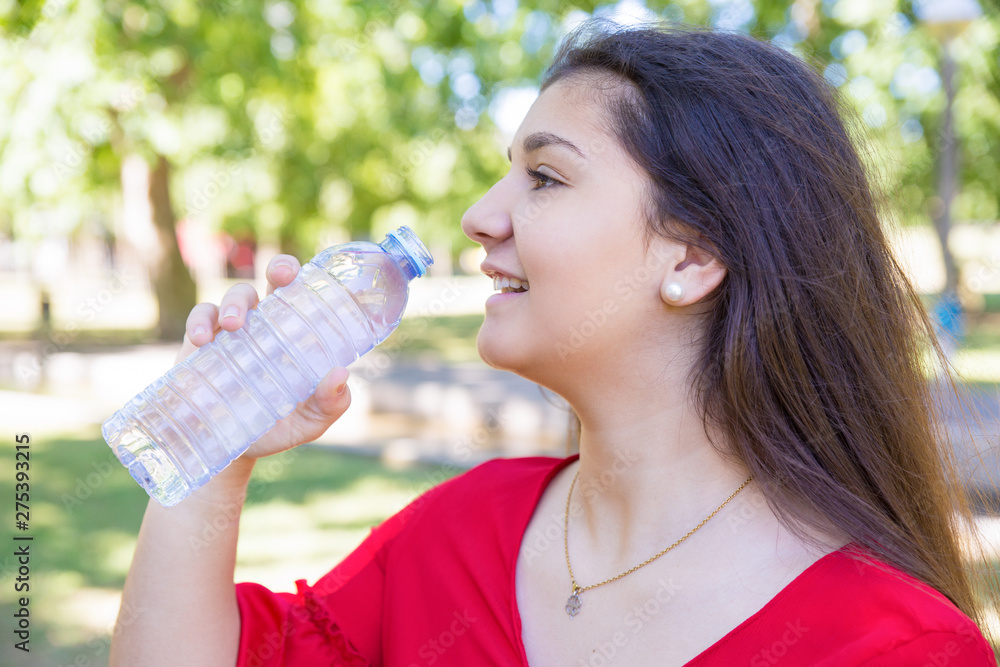Smiling pretty young woman drinking water from bottle in park. Beautiful lady wearing red blouse with blurred green lawn and trees in background. Thirst and nature concept. Side view. Smiling pretty young woman drinking water from bottle in park. Beautiful lady wearing red blouse with blurred green lawn and trees in background. Thirst and nature concept. Side view.