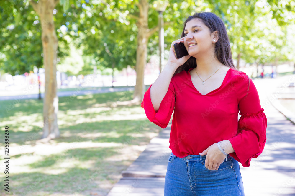 Smiling pretty young woman talking on mobile phone in park. Beautiful lady wearing red blouse and standing with green lawn and trees in background. Communication and nature concept. Front view. Smiling pretty young woman talking on mobile phone in park. Beautiful lady wearing red blouse and standing with green lawn and trees in background. Communication and nature concept. Front view.