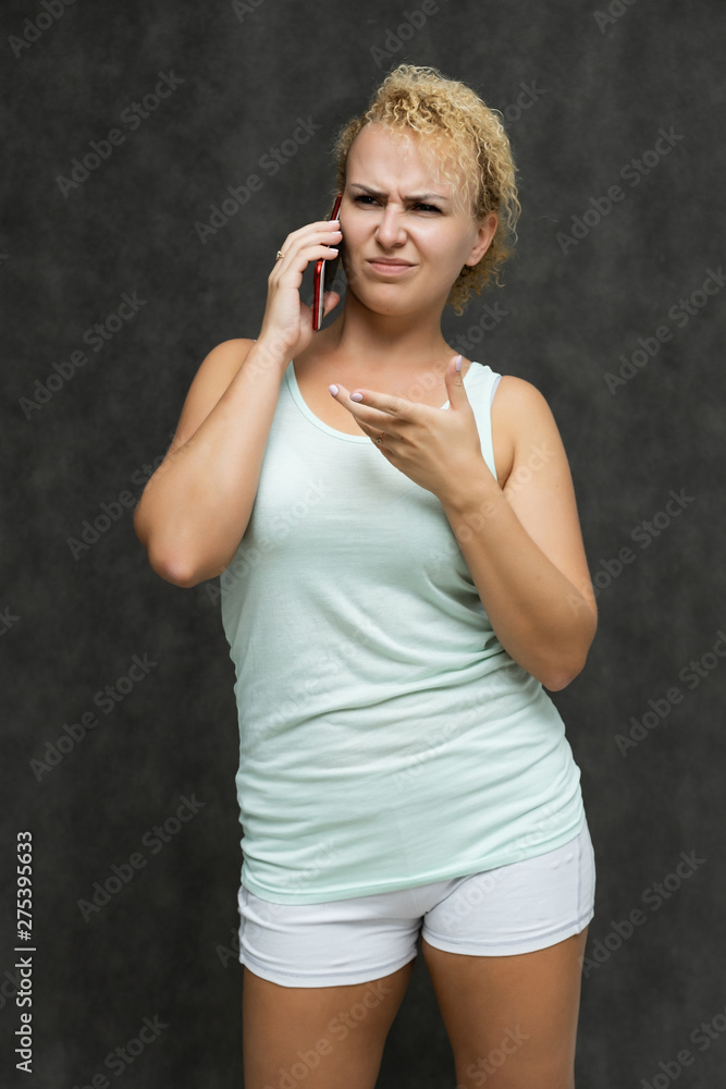 portrait above the knee of a beautiful blonde woman girl with short curly hair on a gray background in bright linen talking on the phone and showing a lot of emotions. Experienced model shows hands. portrait above the knee of a beautiful blonde woman girl with short curly hair on a gray background in bright linen talking on the phone and showing a lot of emotions. Experienced model shows hands.