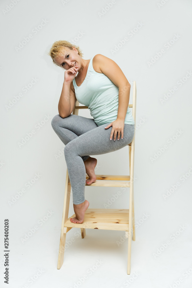 Full-length portrait of a slim beautiful pretty girl blonde woman with short curly hair on a white background in bright fitness clothes sitting on the stairs in various poses and a lot of emotions. Full-length portrait of a slim beautiful pretty girl blonde woman with short curly hair on a white background in bright fitness clothes sitting on the stairs in various poses and a lot of emotions.