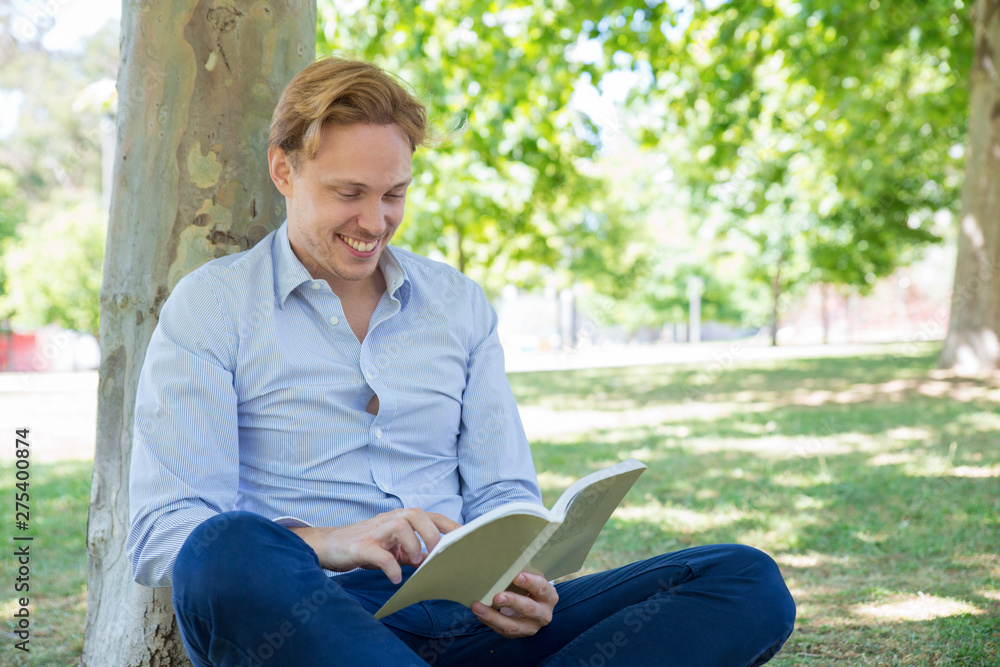 Joyful handsome guy reading in park. Handsome man in shirt sitting on grass, leaning on tree, holding open book and smiling. Happy book reader concept Joyful handsome guy reading in park. Handsome man in shirt sitting on grass, leaning on tree, holding open book and smiling. Happy book reader concept