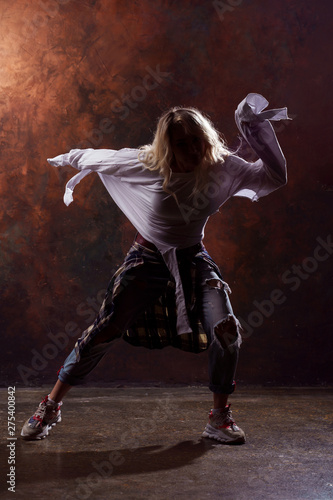 Fototapeta Naklejka Na Ścianę i Meble -  Photo of young dancing blonde girl in ripped jeans and sneakers on dark brown background
