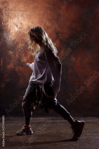 Side photo of slim blonde dancer in torn jeans and sneakers on dark brown background © Sergey