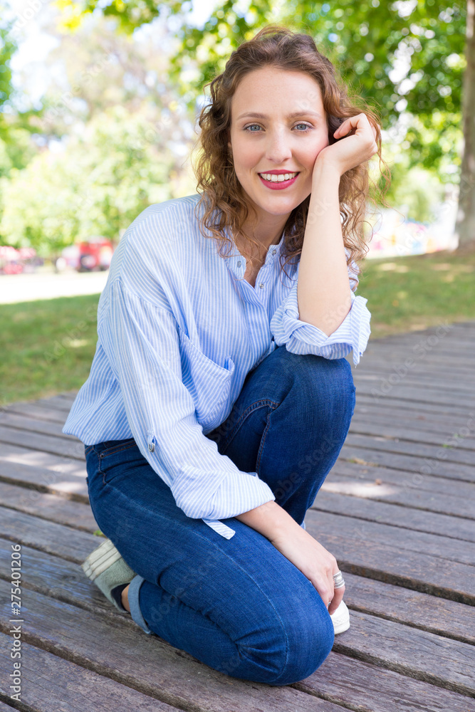 Pensive happy college girl spending leisure time in park. Pretty young woman in casual with wavy hair sitting on walkway and smiling at camera. Weekend outdoors concept Pensive happy college girl spending leisure time in park. Pretty young woman in casual with wavy hair sitting on walkway and smiling at camera. Weekend outdoors concept
