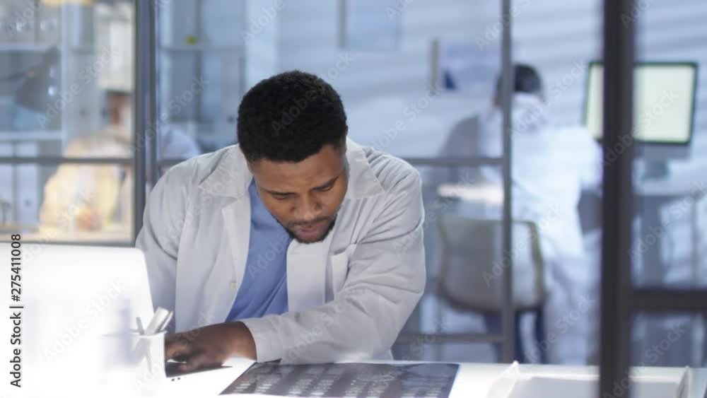 Waist-up shot of concerned Afro-American neurologist, dressed in lab coat and medical uniform, hurriedly studying MRI scan and typing on laptop for patient case record Waist-up shot of concerned Afro-American neurologist, dressed in lab coat and medical uniform, hurriedly studying MRI scan and typing on laptop for patient case record