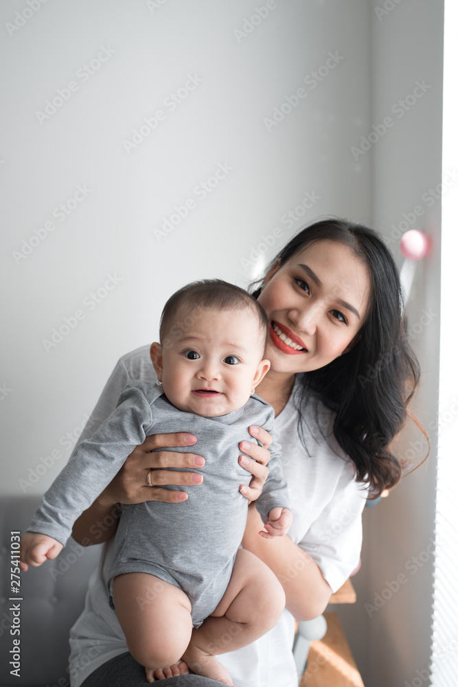 Beautiful young mom is playing with her cute baby and smiling while standing near the window at home Beautiful young mom is playing with her cute baby and smiling while standing near the window at home