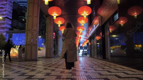 Caucasian girl in a long light raincoat, stands in the middle of the night on Macao street. Nearby are people. Above the girl hang red lanterns with yellow fringe.