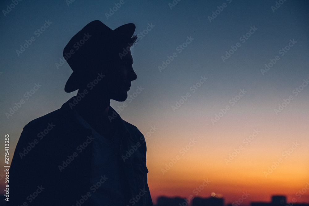 Silhouette of a man's portrait, watching the sunset over the city. Urban man with hat. Good looking