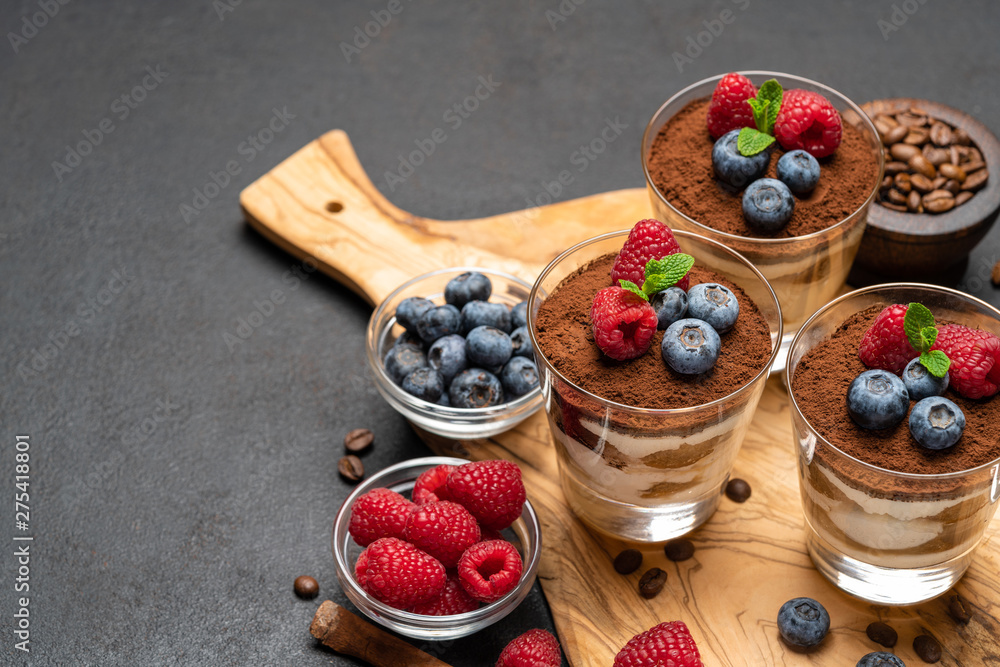 Classic tiramisu dessert with blueberries and raspberries in a glass on wooden cutting board on dark concrete background Classic tiramisu dessert with blueberries and raspberries in a glass on wooden cutting board on dark concrete background