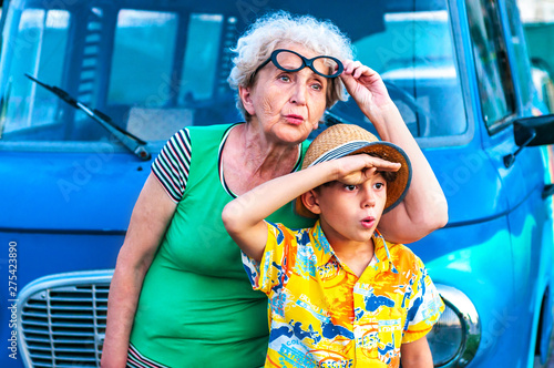 Trendy grandmothers with her grandson in beach colorful clothes near the retro car peer into the distance in surprise. Family summer vacation and travel concept
